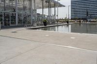 a glass enclosed structure near the water and large buildings in the background with benches on the side, and a small pool, is surrounded by concrete sidewalked area for people sitting and walking around the perimeter