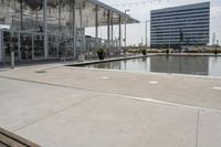 a glass enclosed structure near the water and large buildings in the background with benches on the side, and a small pool, is surrounded by concrete sidewalked area for people sitting and walking around the perimeter