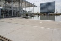 a glass enclosed structure near the water and large buildings in the background with benches on the side, and a small pool, is surrounded by concrete sidewalked area for people sitting and walking around the perimeter