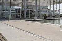 a glass enclosed structure near the water and large buildings in the background with benches on the side, and a small pool, is surrounded by concrete sidewalked area for people sitting and walking around the perimeter