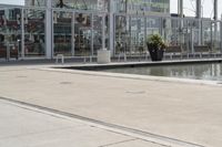 a glass enclosed structure near the water and large buildings in the background with benches on the side, and a small pool, is surrounded by concrete sidewalked area for people sitting and walking around the perimeter