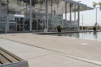 a glass enclosed structure near the water and large buildings in the background with benches on the side, and a small pool, is surrounded by concrete sidewalked area for people sitting and walking around the perimeter