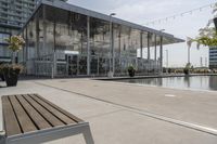 a glass enclosed structure near the water and large buildings in the background with benches on the side, and a small pool, is surrounded by concrete sidewalked area for people sitting and walking around the perimeter