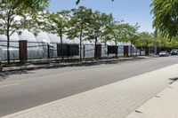 a paved side walk between two trees, a sidewalk and a metal fence along a road