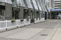 there is a white picketed fence near some tall buildings, a sidewalk and the street is made up from blocks