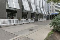 there is a white picketed fence near some tall buildings, a sidewalk and the street is made up from blocks