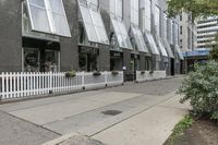 there is a white picketed fence near some tall buildings, a sidewalk and the street is made up from blocks