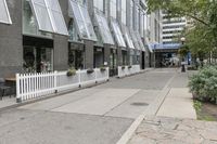 there is a white picketed fence near some tall buildings, a sidewalk and the street is made up from blocks