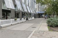 there is a white picketed fence near some tall buildings, a sidewalk and the street is made up from blocks