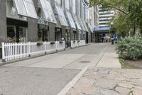 there is a white picketed fence near some tall buildings, a sidewalk and the street is made up from blocks