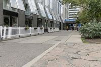 there is a white picketed fence near some tall buildings, a sidewalk and the street is made up from blocks