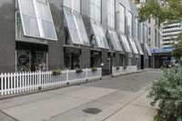 there is a white picketed fence near some tall buildings, a sidewalk and the street is made up from blocks