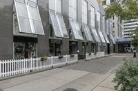 there is a white picketed fence near some tall buildings, a sidewalk and the street is made up from blocks