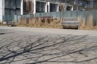 a empty park bench in front of tall buildings with tall grass in the background and an empty sidewalk behind