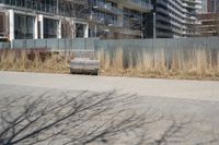 a empty park bench in front of tall buildings with tall grass in the background and an empty sidewalk behind