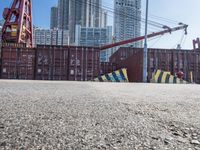 a crane near a long metal fence on top of a road with shipping boxes in the background