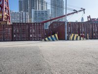 a crane near a long metal fence on top of a road with shipping boxes in the background