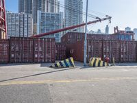 a crane near a long metal fence on top of a road with shipping boxes in the background