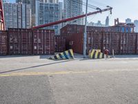 a crane near a long metal fence on top of a road with shipping boxes in the background