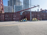 a crane near a long metal fence on top of a road with shipping boxes in the background