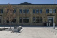 large brick building with a clock sitting in front of it and a tree sitting on the sidewalk