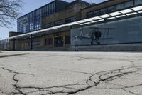 the ground in front of this empty building is cracked and some people are walking and waiting at the bus stop