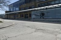 the ground in front of this empty building is cracked and some people are walking and waiting at the bus stop