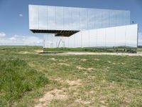 an unusual, mirrored sculpture stands alone in a grassy field under a blue sky with clouds