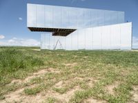 an unusual, mirrored sculpture stands alone in a grassy field under a blue sky with clouds