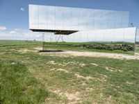 an unusual, mirrored sculpture stands alone in a grassy field under a blue sky with clouds