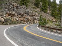 the curved road curves in front of a mountain area with pine trees behind it,