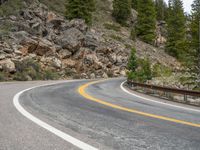 the curved road curves in front of a mountain area with pine trees behind it,