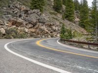 the curved road curves in front of a mountain area with pine trees behind it,