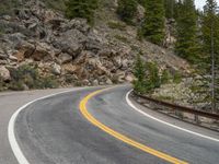 the curved road curves in front of a mountain area with pine trees behind it,