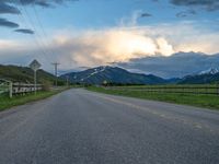 the road is empty during the day and has mountains in the distance as well as a fence