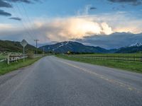 the road is empty during the day and has mountains in the distance as well as a fence