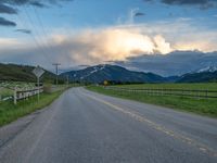 the road is empty during the day and has mountains in the distance as well as a fence