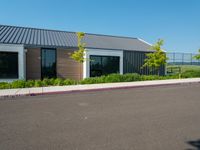 an asphalted parking lot in front of a building and several trees, bushes, and a fence