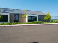 an asphalted parking lot in front of a building and several trees, bushes, and a fence