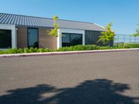 an asphalted parking lot in front of a building and several trees, bushes, and a fence