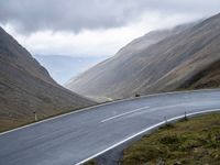 the curved asphalt road is flanked by large mountains and cloudy skies with clouds in the sky