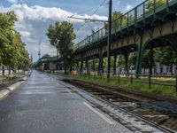 a train traveling down a train track next to a bridge on a street side in front of trees and sky