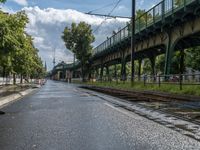 a train traveling down a train track next to a bridge on a street side in front of trees and sky