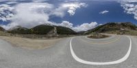 the panoramic shot shows an empty roadway near a mountainous area with a steep hill and blue sky