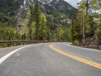 the curved road curves in front of a mountain area with pine trees behind it,