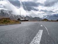 the empty highway is near mountains under a cloudy sky above a small cabin in a valley