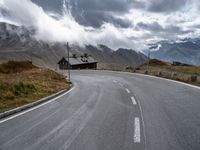 the empty highway is near mountains under a cloudy sky above a small cabin in a valley