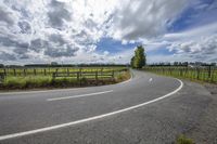 an empty paved road in front of a vineyard field and fenced in area with wooden posts