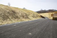 an asphalt road with some rocks near it and trees in the background, with hills and trees to the right, on one side of it