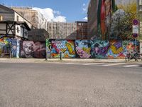 a wall with different colorful artwork on it in a city with buildings around and the street has a bike path going under it